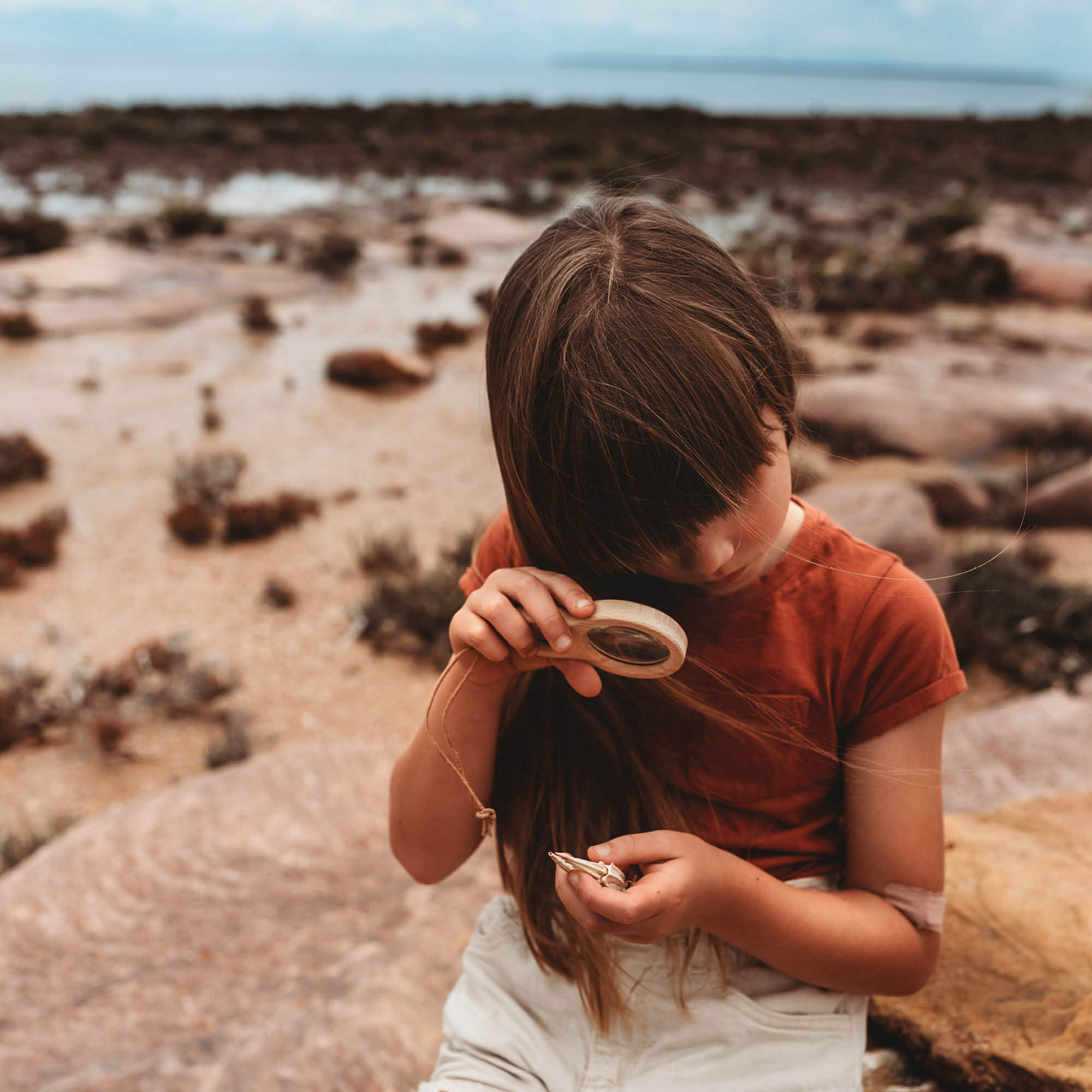 Girl at beach looking at crab claw with a Kikkerland dual magnifier wooden magnifying glass with two lenses for exploring nature and play from Your Wild Books