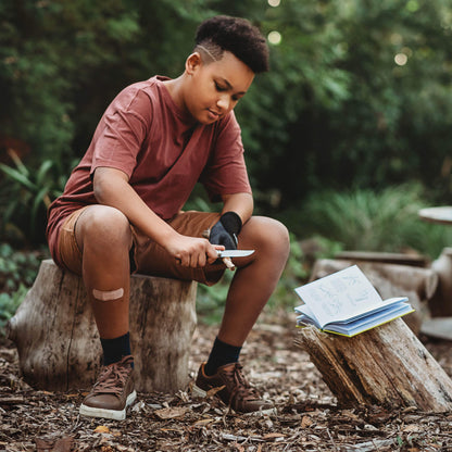Boy sitting in nature with whittling knife and book 50 things to do with a penknife book wood whittling book for kids wood working projects from Your Wild Books
