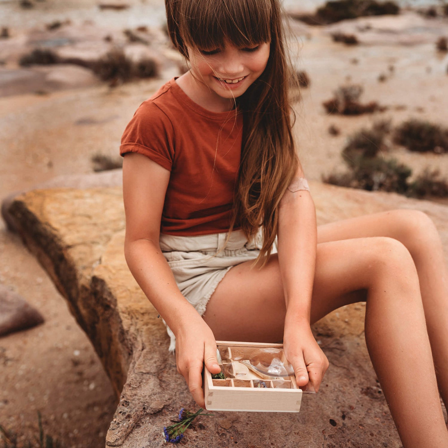 Girl at beach placing nature inside Wooden bug box made by Kikkerland from Your Wild Books. Crab claw, flowers, and shells inside the wooden compartments with magnifying lenses.