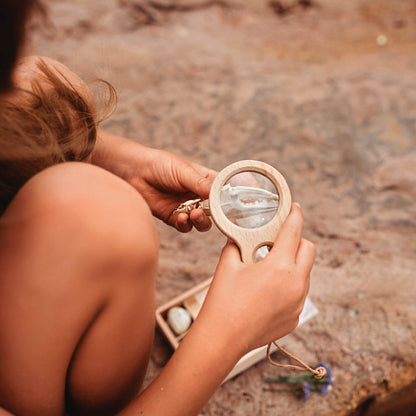 Girl at beach looking at crab claw with a Kikkerland dual magnifier wooden magnifying glass with two lenses for exploring nature and play  from Your Wild Books