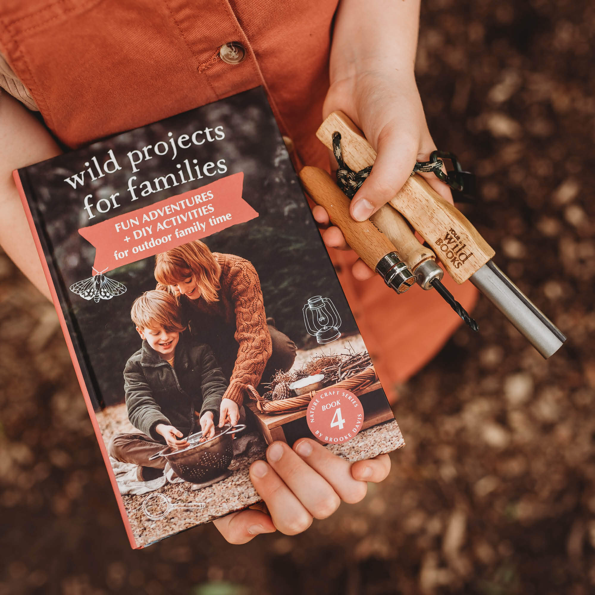 Child holding the Real Tools Bundle by Your Wild Books includes Wild Projects for Families book, whittling knife, hand drill and fire starter.
