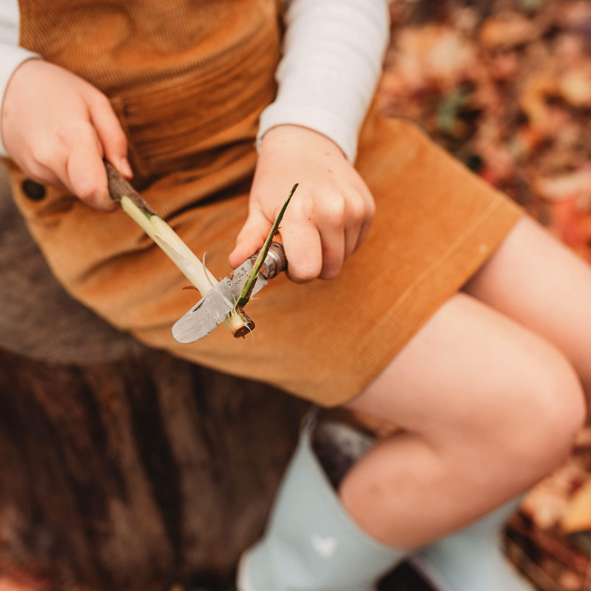Child sitting in woods peeling bark from a stick. Wood whittling knives for kids for all your nature craft projects, suitable for beginners and advanced woodworkers. Made by Opinel from Your Wild Books.