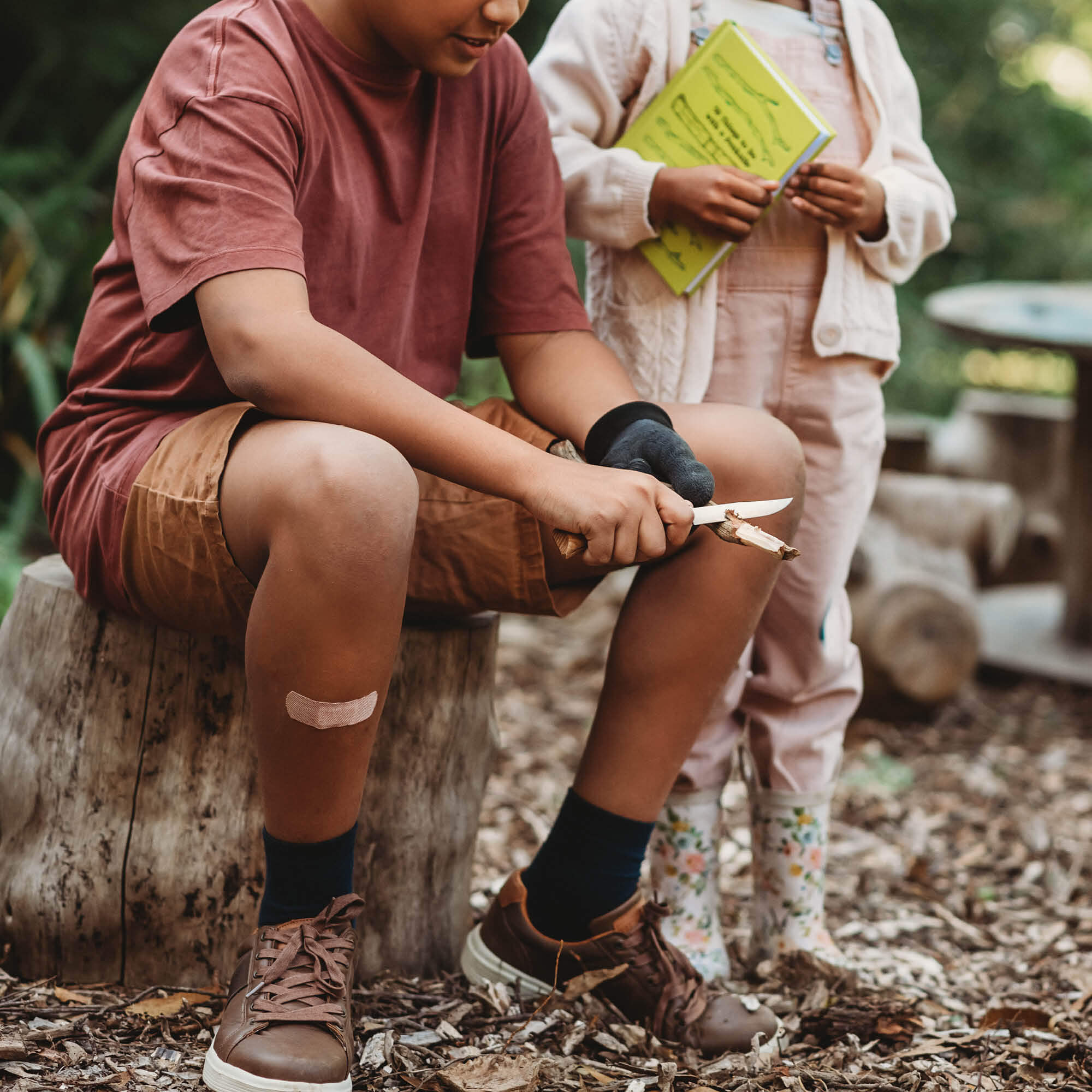 Child sitting in woods peeling bark from a stick. Wood whittling knives for kids for all your nature craft projects, suitable for beginners and advanced woodworkers. Made by Opinel from Your Wild Books.