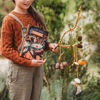 Child holding a wind catcher made from nature with hanging gum nuts, shells, flowers and seed pods. Step by step instructions are found in Wild Child, nature craft projects for kids book, made in Australia by Your Wild Books. 