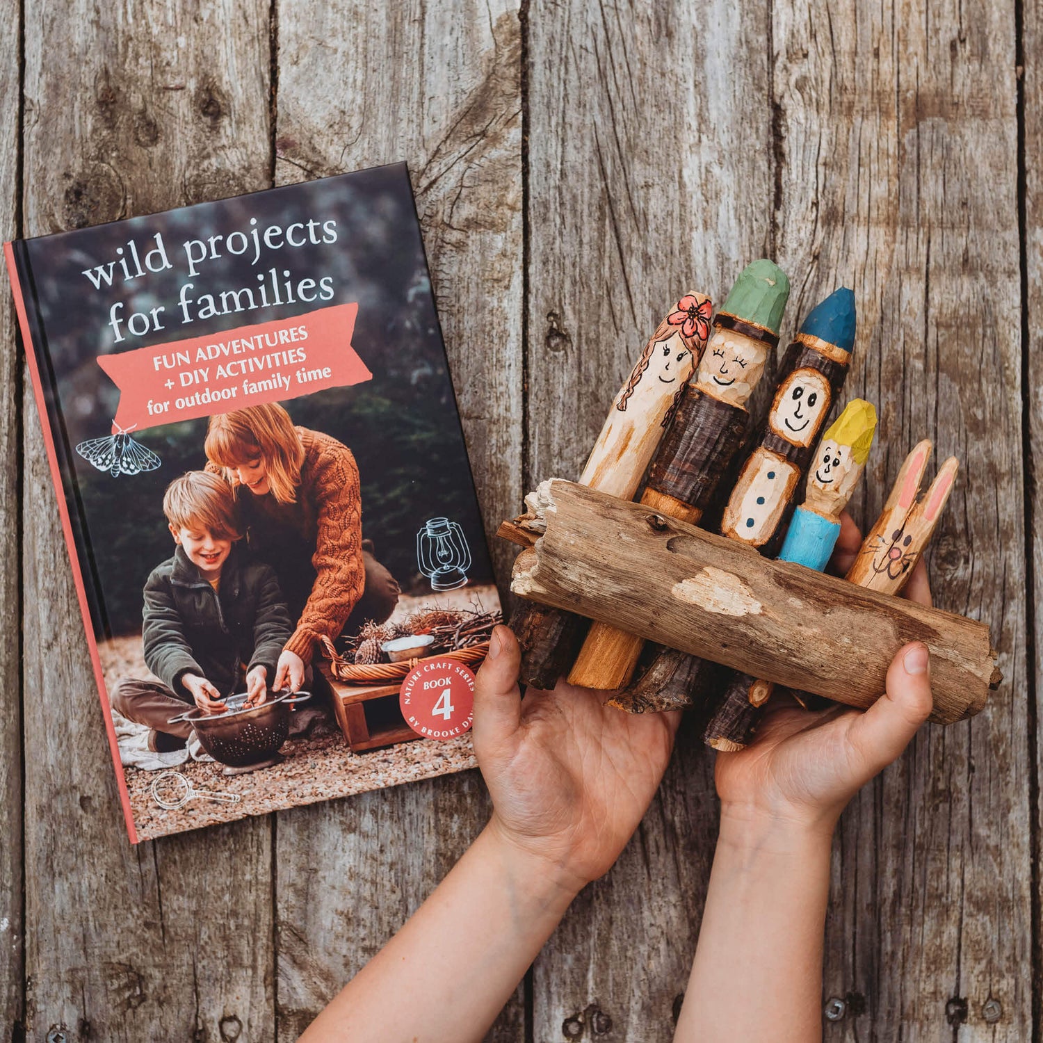 Child holding a whittled stick family decorated with paint pens next to the book Wild Projects for Families from the Nature Craft Series by Your Wild Books is printed in Australia using FSC Certified paper.